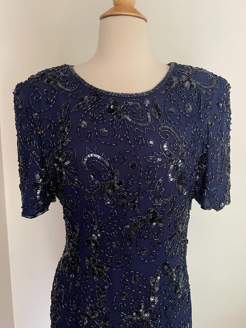 May include: Navy blue dress with short sleeves, embellished with intricate black sequins and beads. The dress features a round neckline and a detailed floral pattern across the bodice and sleeves. The dress is displayed on a mannequin.