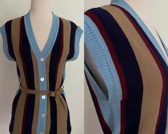 May include: A sleeveless, button-down sweater vest with vertical stripes in navy, tan, burgundy, and light blue. The vest has a V-neck and a matching belt. The buttons are pearlescent. The vest is displayed on a mannequin.