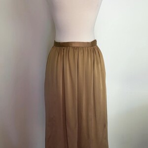 May include: A midi-length skirt in a solid bronze-brown color with a satin-like finish. The skirt features a smooth, flat waistband and a gathered, pleated design that creates a flowing silhouette. The fabric has a subtle sheen throughout.
