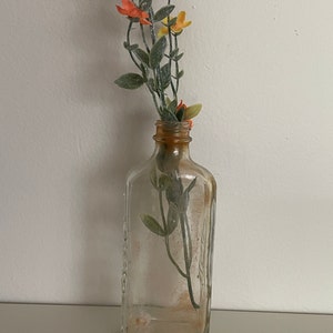 Könnte beinhalten: Eine klare, rechteckige Glasflasche mit braunem Hals, die einen Zweig künstlicher Blumen enthält. Die Blumen haben orange und gelbe Blütenblätter mit grünen Blättern. Die Flasche steht auf einer weißen Oberfläche.