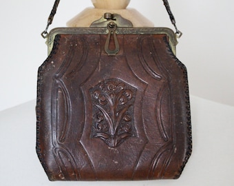 May include: A vintage brown leather handbag with a decorative floral design. The bag features a metal clasp and a stitched leather strap. The leather has a textured appearance, and the bag is likely from the early 20th century.