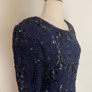 May include: A navy blue dress with short sleeves, embellished with intricate black beading and sequins. The dress features a round neckline and a detailed floral pattern, creating a formal and elegant look. The dress is displayed on a mannequin.