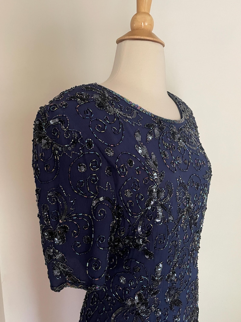 May include: A navy blue dress with short sleeves, embellished with intricate black beading and sequins. The dress features a round neckline and a detailed floral pattern, creating a formal and elegant look. The dress is displayed on a mannequin.