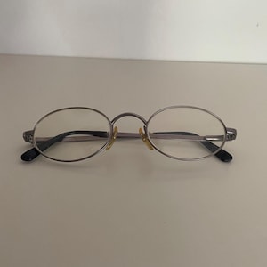 May include: Oval-shaped eyeglasses with a silver-colored frame and black temples. The lenses are clear, and the bridge of the glasses is a matching silver color. The glasses are designed for vision correction.