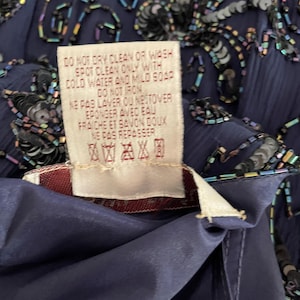 May include: Close-up of a navy blue garment with intricate beadwork and sequins. A care label is visible, with text indicating spot cleaning instructions. The fabric appears to be a flowing, possibly silk-like material.