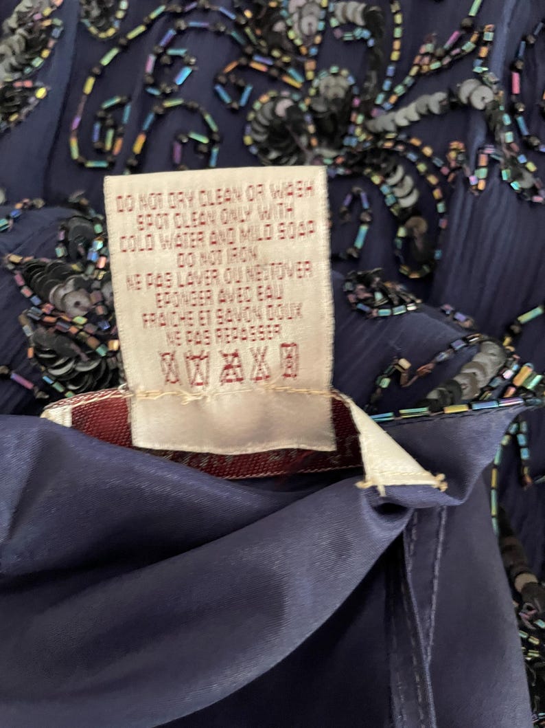 May include: Close-up of a navy blue garment with intricate beadwork and sequins. A care label is visible, with text indicating spot cleaning instructions. The fabric appears to be a flowing, possibly silk-like material.