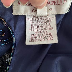 May include: Close-up of a navy blue garment tag with the brand name "A. MAIA CAPELL" and detailed fabric composition information. The tag is held by a hand, showing the garment's shell is 100% silk and the lining is 100% polyester.