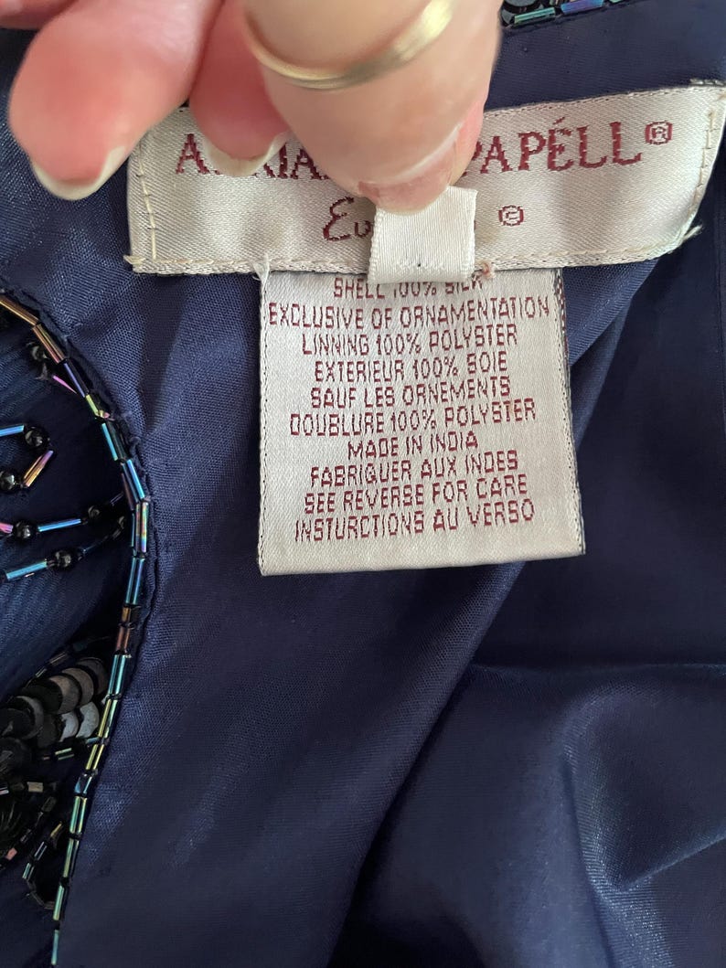 May include: Close-up of a navy blue garment tag with the brand name "A. MAIA CAPELL" and detailed fabric composition information. The tag is held by a hand, showing the garment's shell is 100% silk and the lining is 100% polyester.