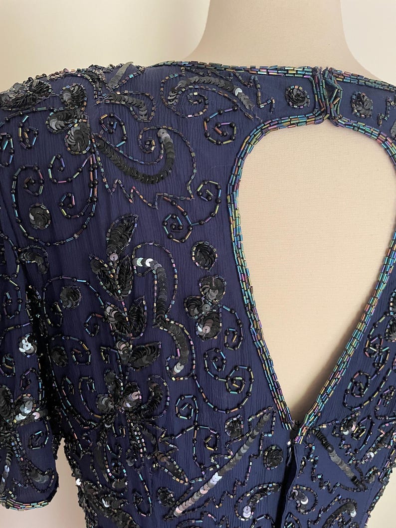 May include: Navy blue formal dress with intricate sequin and bead embellishments. The dress features a keyhole back and short sleeves. The design includes floral and swirling patterns, with iridescent accents around the neckline and keyhole.