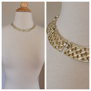May include: A gold-toned necklace with a woven design is displayed on a mannequin. The necklace has a collar style with a clasp closure. The woven pattern is visible in the close-up shot, showcasing the intricate details of the jewelry.