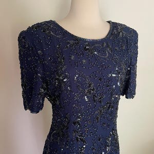 May include: A navy blue dress with short sleeves, adorned with intricate black sequin and bead embellishments. The dress features a round neckline and a fitted silhouette, showcasing a floral pattern. The dress is displayed on a mannequin.