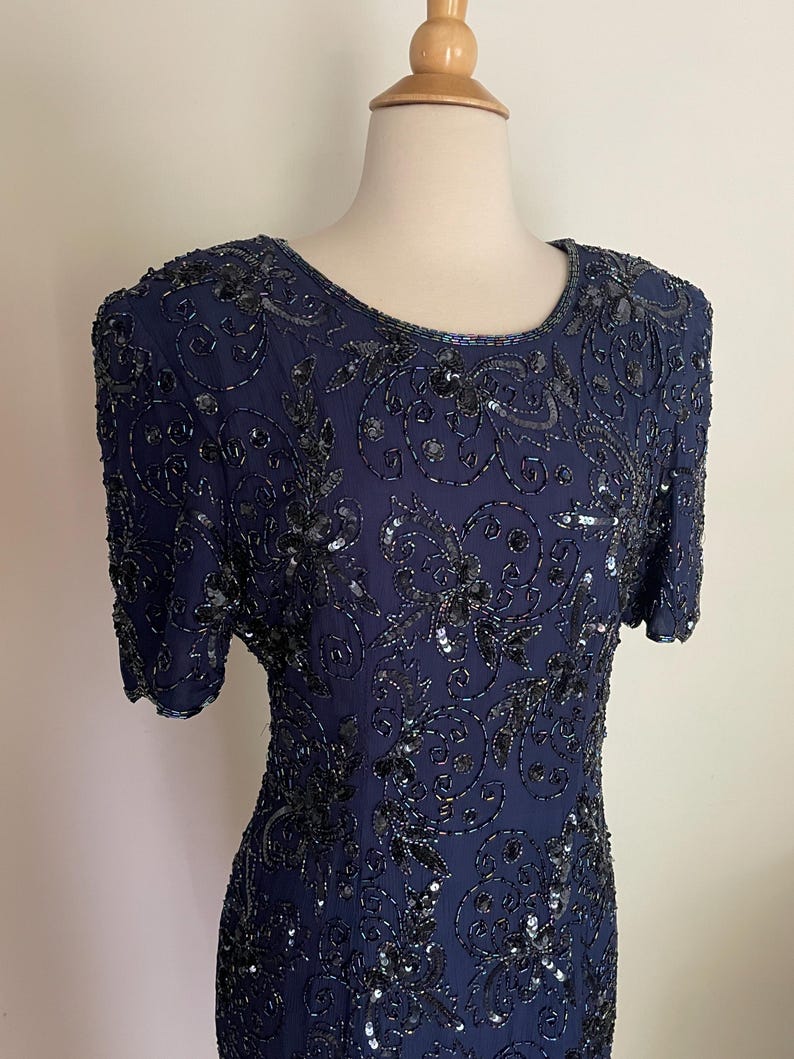 May include: A navy blue dress with short sleeves, adorned with intricate black sequin and bead embellishments. The dress features a round neckline and a fitted silhouette, showcasing a floral pattern. The dress is displayed on a mannequin.