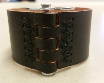May include: Black leather cuff bracelet with a metal clasp and decorative stitching. The stitching is in a braided pattern.