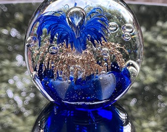 May include: A spherical glass paperweight featuring a clear exterior with suspended air bubbles. Inside, a deep blue floral-like structure is surrounded by shimmering gold-colored metallic flecks, all set against a dark blue base.