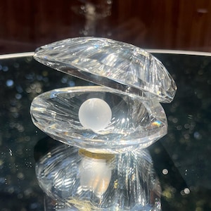 May include: A clear, crystal clam shell sculpture with an open top, revealing a white pearl. The shell has a textured, faceted design. The sculpture sits on a reflective surface, with a blurred background.