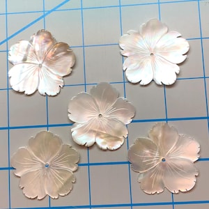 May include: Five iridescent, mother-of-pearl flower-shaped embellishments. Each flower has five petals and a small hole in the center. The flowers are white with subtle pink and yellow hues, and are arranged on a grid.