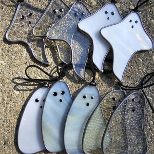 May include: A set of nine stained glass ghost ornaments. The ghosts are in various shades of white, grey, and clear. Some of the ghosts have a textured surface, while others are smooth. The ghosts are all hanging from black cords.