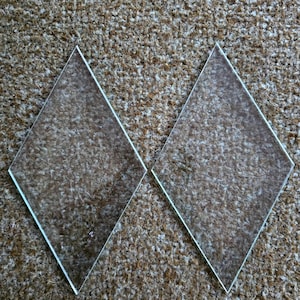 May include: Two clear, diamond-shaped glass pieces with beveled edges. The glass is transparent, allowing the background to be visible. The pieces are positioned on a textured, brown surface.
