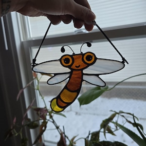 May include: A stained glass dragonfly suncatcher with a whimsical design. The dragonfly features a yellow, orange, and brown body with large, round eyes and clear wings. It is suspended by black cords, perfect for hanging in a window.