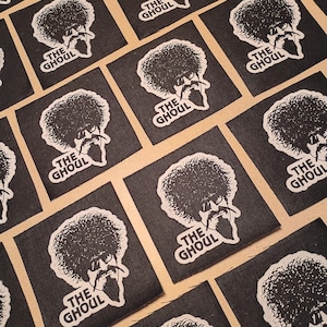 May include: A collection of square black fabric patches, each featuring a white screen-printed graphic of a person with a large afro hairstyle, sunglasses, and a goatee. Below the illustration, the text THE GHOUL is printed in white capital letters.