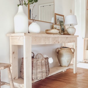 May include: A light wood console table with a white vase, a white ceramic vase, a woven basket, and a large ceramic vase. The table has a shelf underneath and is sitting on a wood floor.