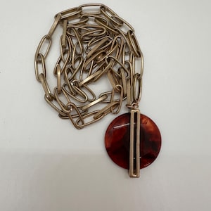 Pode incluir: Um colar de corrente de ouro com um pingente redondo vermelho e marrom. O pingente tem uma barra de ouro no centro.