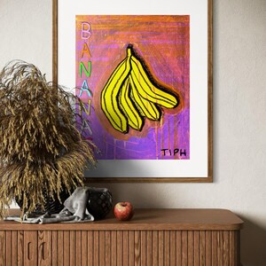 Pode incluir: Uma impressão emoldurada de uma pintura de um cacho de bananas amarelas sobre um fundo roxo e laranja. A palavra "BANANA" está escrita verticalmente em letras coloridas no lado esquerdo da pintura. A assinatura do artista "TIPH" está escrita em preto no canto inferior direito da pintura.