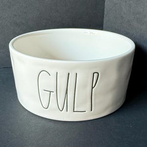 May include: A white ceramic bowl with a slightly uneven, organic texture. The word GULP is debossed in black, thin, handwritten-style lettering on the front exterior of the bowl. The interior is smooth and white, featuring a wide, open circular rim.
