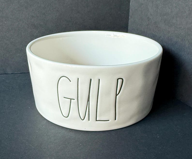 May include: A white ceramic bowl with a slightly uneven, organic texture. The word GULP is debossed in black, thin, handwritten-style lettering on the front exterior of the bowl. The interior is smooth and white, featuring a wide, open circular rim.