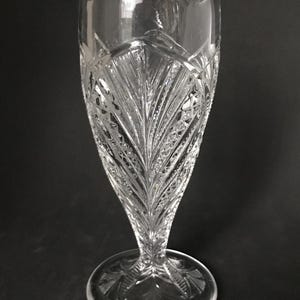May include: Clear, cut-glass vase with a flared top and a round base. The vase features a detailed, etched pattern of leaves and geometric shapes. The design is symmetrical, with a tall, slender profile. The vase is approximately 10 inches tall.