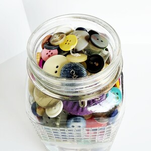 May include: A clear glass jar filled with a variety of colorful buttons. The buttons are in various sizes, shapes, and colors, including yellow, black, white, and blue. The jar is sitting on a white surface.