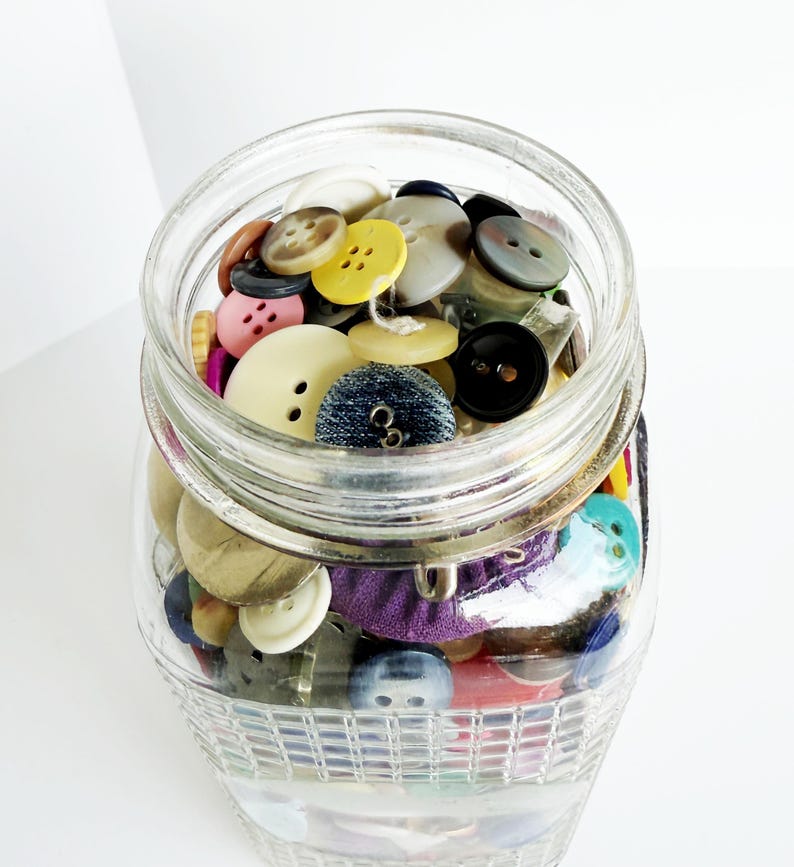 May include: A clear glass jar filled with a variety of colorful buttons. The buttons are in various sizes, shapes, and colors, including yellow, black, white, and blue. The jar is sitting on a white surface.