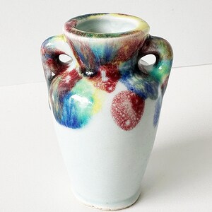 May include: A small, ceramic vase with two handles. The vase has a white base and is decorated with colorful, speckled paint in shades of red, blue, yellow, and green. The rim of the vase is yellow.