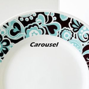 May include: White ceramic plate with a decorative border featuring a floral design in black and teal. The word "Carousel" is printed on the plate.