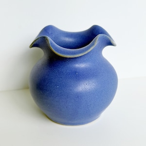 May include: A small, rounded ceramic vase with a vibrant blue matte finish. The vessel features a distinctively wide, ruffled, and wavy rim that curves outward, creating a soft, undulating edge. The surface has a subtle, uniform texture.