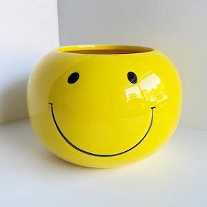 May include: A round, yellow ceramic planter with a smiley face design. The planter features black eyes and a curved black line for a mouth. The glossy finish reflects light, enhancing the cheerful design. A decorative item for home or garden.