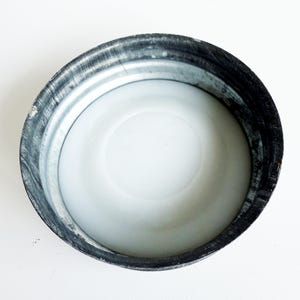 May include: A close-up view of a metal lid with a white interior. The lid has a silver-colored exterior with a dark, textured rim. The interior is smooth and white, creating a contrast with the metallic outer edge.