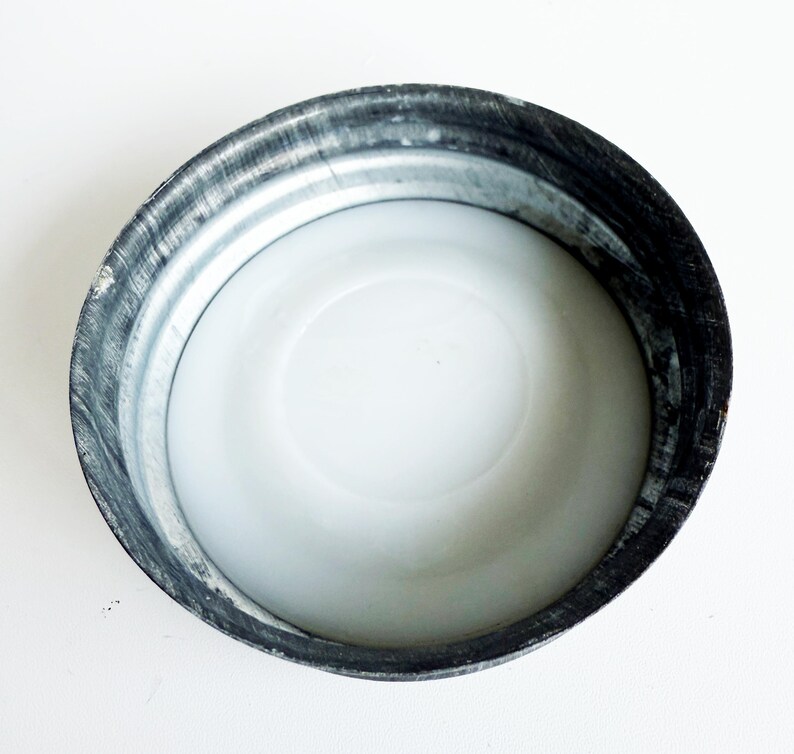 May include: A close-up view of a metal lid with a white interior. The lid has a silver-colored exterior with a dark, textured rim. The interior is smooth and white, creating a contrast with the metallic outer edge.