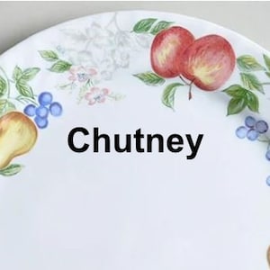May include: White ceramic dinner plate with a floral design featuring pears, apples, and blueberries. The plate has the word "Chutney" written in black lettering.