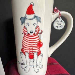 May include: White ceramic mug with a Christmas dog design. The dog is wearing a red and white striped sweater, a Santa hat, and holding a candy cane. The mug has a tag that says "fetch the spirit".