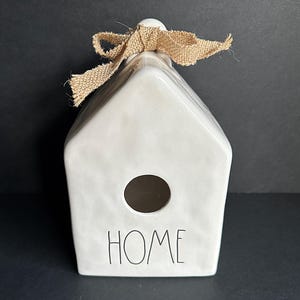 May include: A white ceramic house-shaped ornament with a textured surface, featuring a circular opening in the center and the word HOME printed in black lettering. A tan burlap ribbon is tied in a bow at the top of the peaked roof structure.