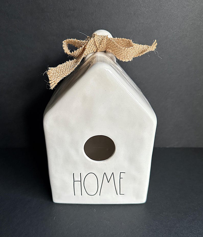 May include: A white ceramic house-shaped ornament with a textured surface, featuring a circular opening in the center and the word HOME printed in black lettering. A tan burlap ribbon is tied in a bow at the top of the peaked roof structure.