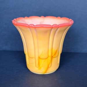 May include: A fluted glass vase featuring a translucent orange and yellow marbled pattern with a vibrant coral-pink scalloped rim. The vessel has a wide, open top that tapers down to a circular base, characterized by vertical ridges along the exterior.