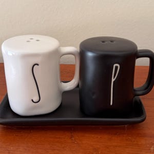 May include: A white ceramic salt shaker and a black ceramic pepper shaker, both shaped like small mugs with handles. The white shaker features a black letter S, and the black shaker features a white letter P. They sit on a black rectangular tray.
