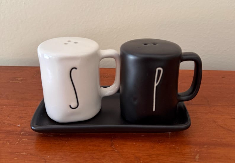 May include: A white ceramic salt shaker and a black ceramic pepper shaker, both shaped like small mugs with handles. The white shaker features a black letter S, and the black shaker features a white letter P. They sit on a black rectangular tray.