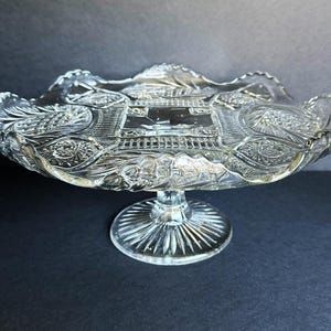 May include: Clear glass pedestal bowl with a scalloped rim and intricate embossed design. The bowl features a detailed pattern of architectural elements and floral motifs, supported by a round, ribbed base. A decorative piece for serving or display.