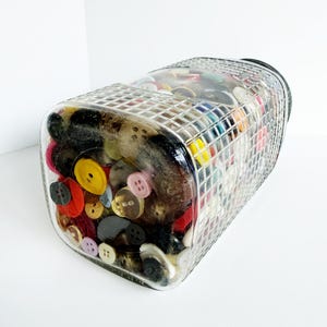 May include: A clear, rectangular glass jar filled with a variety of colorful buttons. The buttons are in various sizes, shapes, and colors, including red, yellow, black, and pink. The jar has a grid-like pattern on the top.