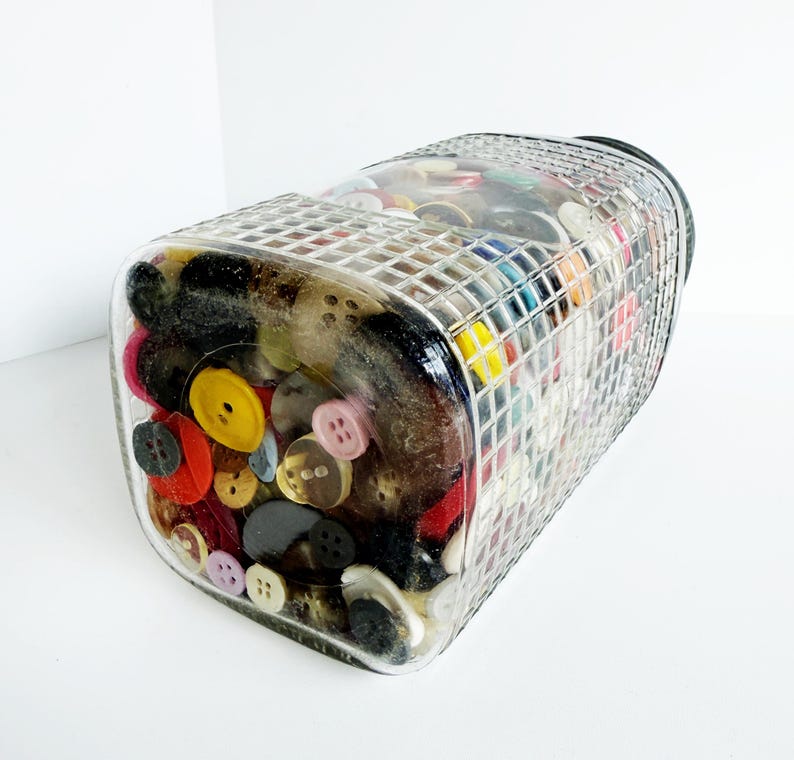 May include: A clear, rectangular glass jar filled with a variety of colorful buttons. The buttons are in various sizes, shapes, and colors, including red, yellow, black, and pink. The jar has a grid-like pattern on the top.