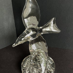 May include: A clear glass sculpture of a bird in flight. The bird is perched on a round, textured base.
