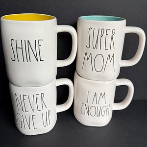 May include: Four white ceramic mugs with black lettering. The top left mug has a yellow interior and says SHINE. The bottom left mug says NEVER GIVE UP. The top right mug has a teal interior and says SUPER MOM. The bottom right mug says I AM ENOUGH.